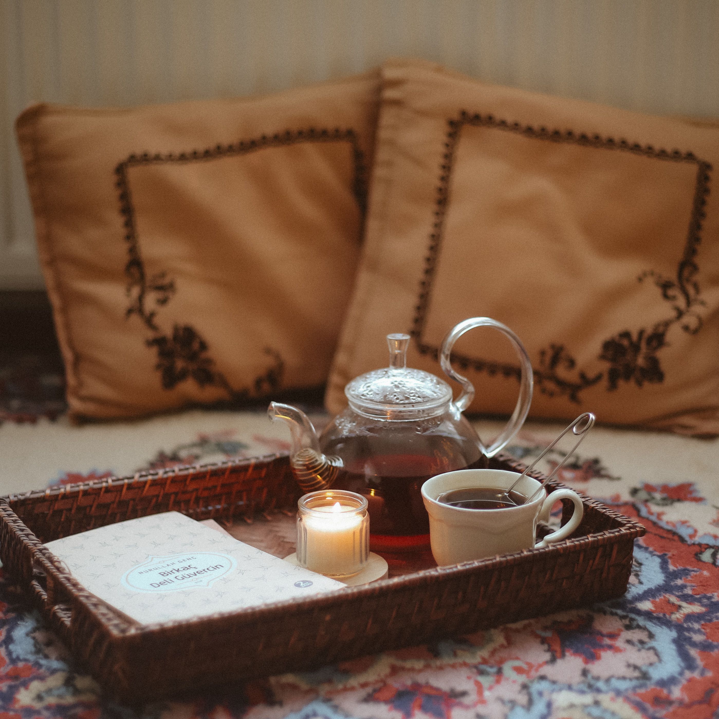 Glass teapot and cup on a woven tray with a candle and cushions
